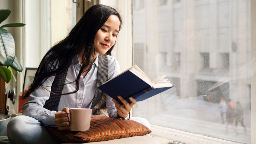 Young asian woman sitting by the window on the sofa with a cup of tea, book and pillow, laughing. Remote work at home. 