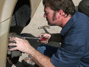 Mechanic crouching down next to a car, holding a screwdriver.