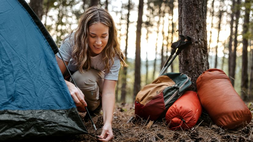 Beautiful smiling women setting up tent in forest with backpack resting near tree