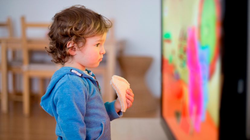 Todler is very close to the tv watching cartoons with a sandwich in his hand