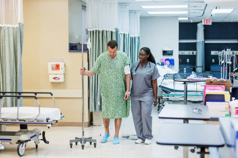 A male patient in a hospital gown with IV drip, walking, helped by a nurse in scrubs. The patient is a mature man in his 50s and the healthcare worker is a young African American woman in her 20s.