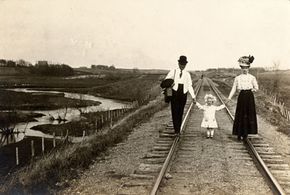 A husband and wife walk on railroad tracks as each holds a hand of their young daughter. Trains made long distance travel much faster and more comfortable and were the preferred way to get around before cars were introduced.