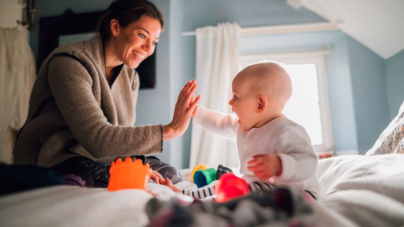 Baby girl is sitting on a double bed with her mother and they have baby toys and stacking cups around hem. The little girl is high-fiving her mother.