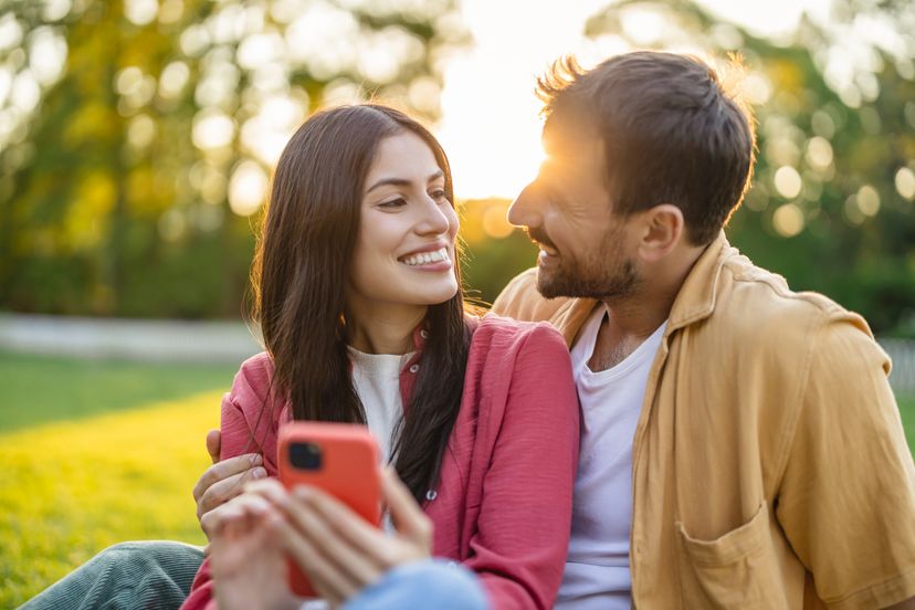 The couple, sitting closely together on a picnic blanket, shares a moment of happiness and smiles while enjoying their time outdoors in a sunlit park with vibrant greenery.