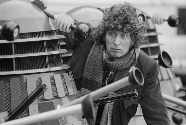 A black-and-white image of actor Tom Baker crouching behind a robot.