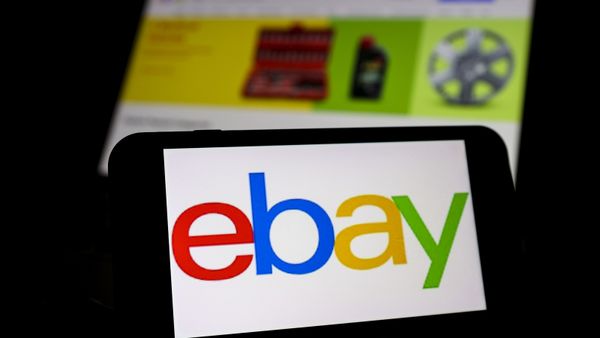 The logo of eBay is displayed on a smartphone and the website of eBay is displayed on a screen behind it