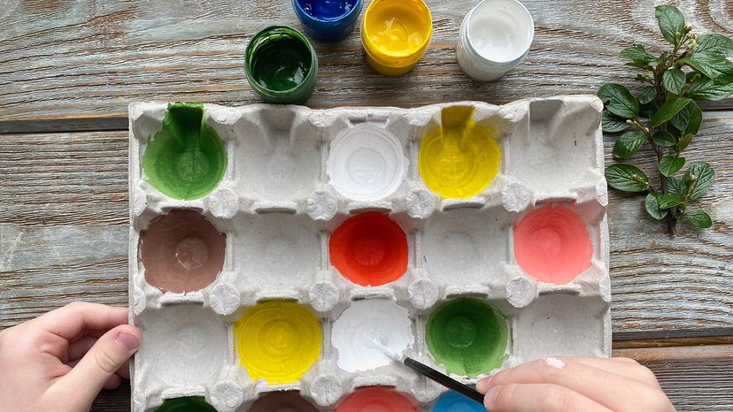 Children's hands paint colored egg box on a wooden background.