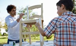 A man and a young boy paint a wooden chair with white paint outdoors.