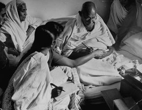 Indian leader Mohandas K. Gandhi, sitting on a mattress on the floor, surrounded by people at the end of his last fast.