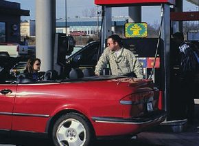 Man filling the gas tank of a red car while talking to woman in passenger seat.