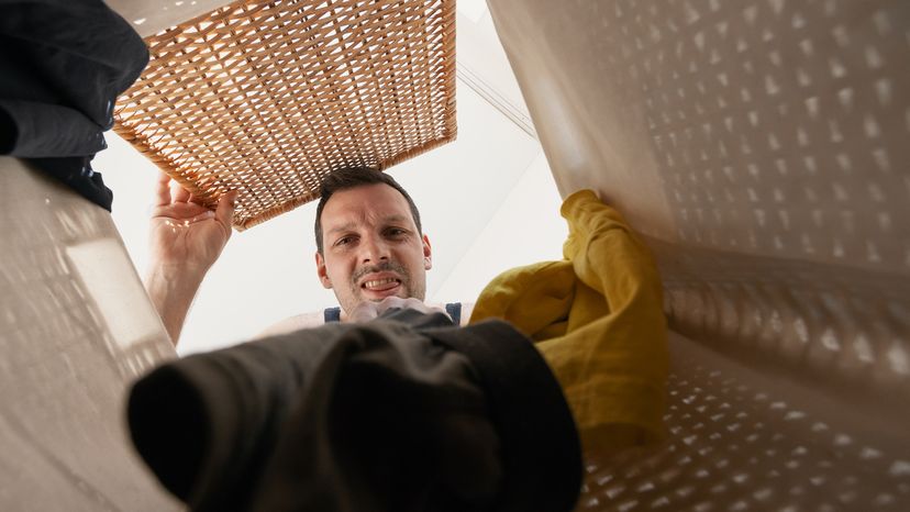 An unshaven man grimaces as he looks into a basket of dirty laundry.