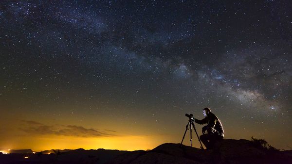 View of Milky Way against photographer with camera sitting on rock.