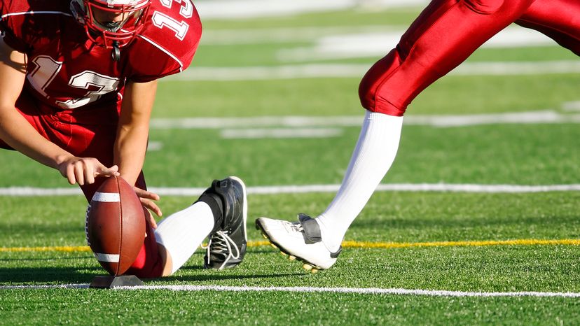 A football player tees up the ball for the approaching kicker, both on red jerseys