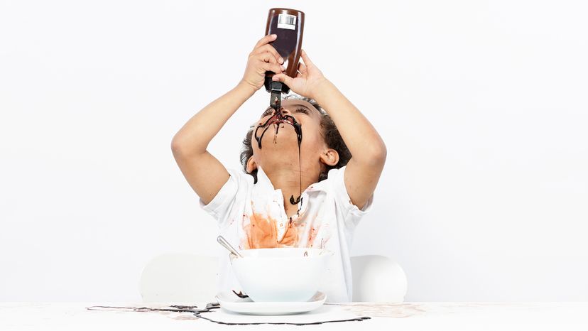 Child in stained white shirt pouring chocolate syrup on his face and clothes