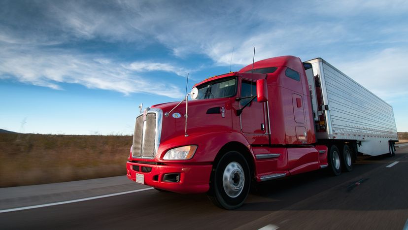 Red semi truck with a white trailer driving down highway on a sunny day