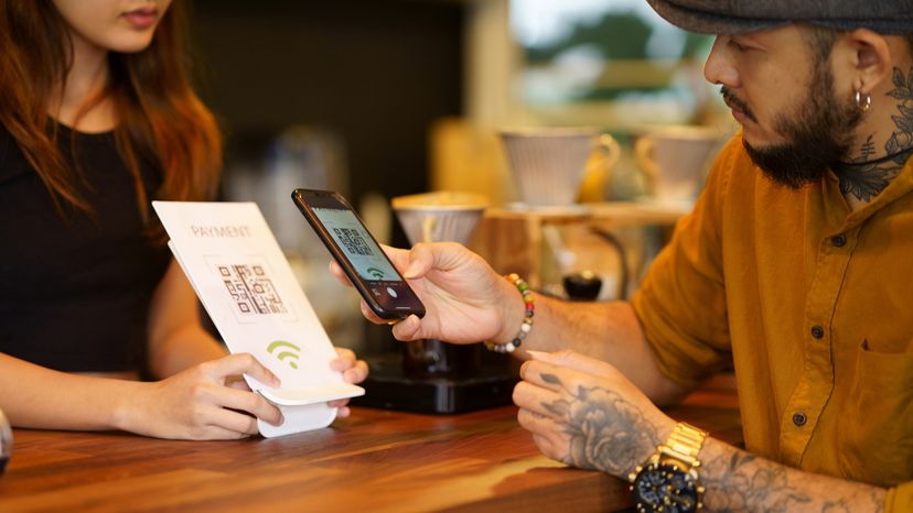 A woman holds a card with a QR code for a man to scan so he can pay for his coffee