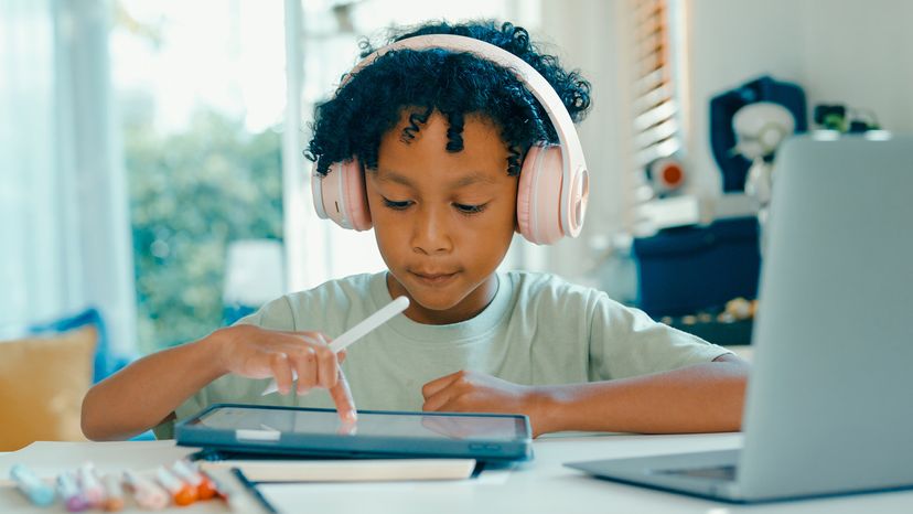 Child wearing pink headphones, holding a stylus and using a tablet at a home desk