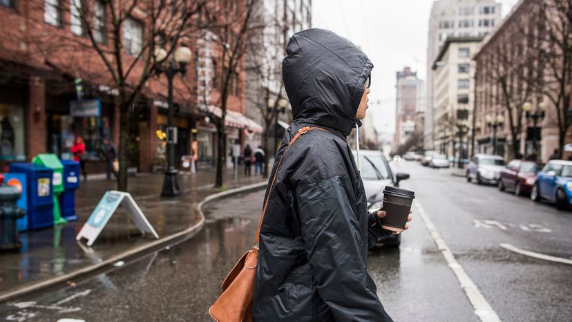 Person walking across a rainy street with their hood up and holding a coffee cup