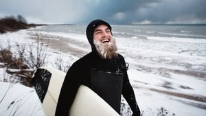 man with icicles in his beard holding a surfboard on snowy shores