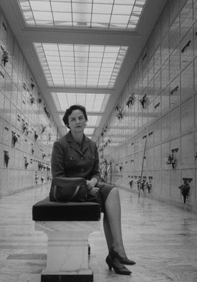 Black-and-white photo of a woman, Jessica Mitford, sitting in a burial vault.