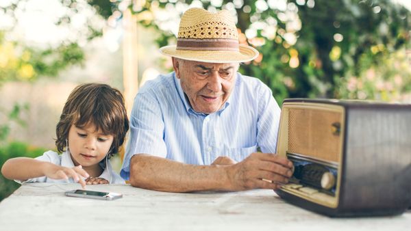 Small kid listening music from a smart phone while his grandfather from an old radio