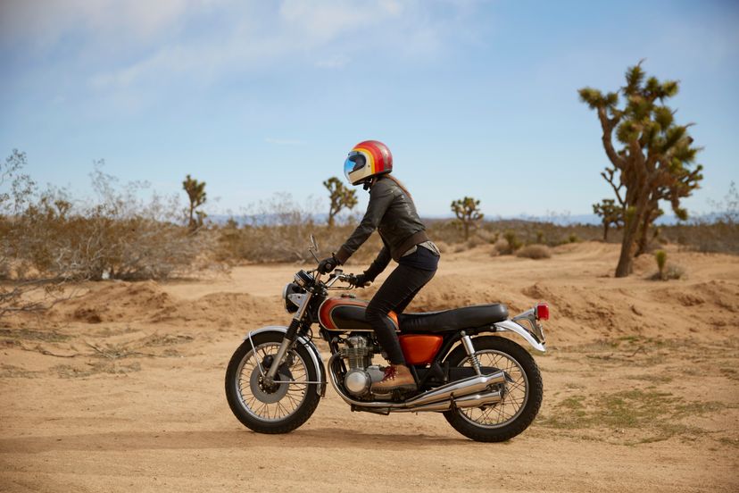 A slim figure in black clothes and a red helmet rides a red motorcycle through desert terrain.
