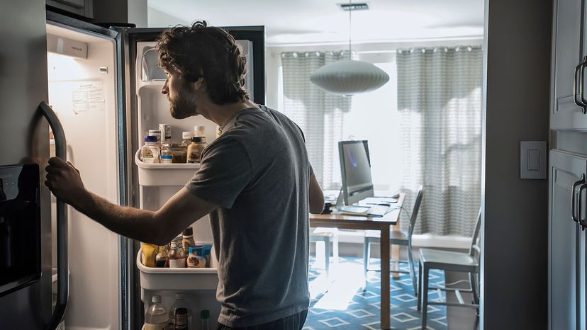 Man in pajamas looks in open refrigerator. Desktop computer is on kitchen table behind him.