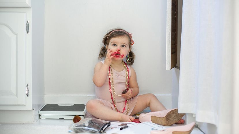 A young girl sits on a bathroom floor with lipstick smeared on her face