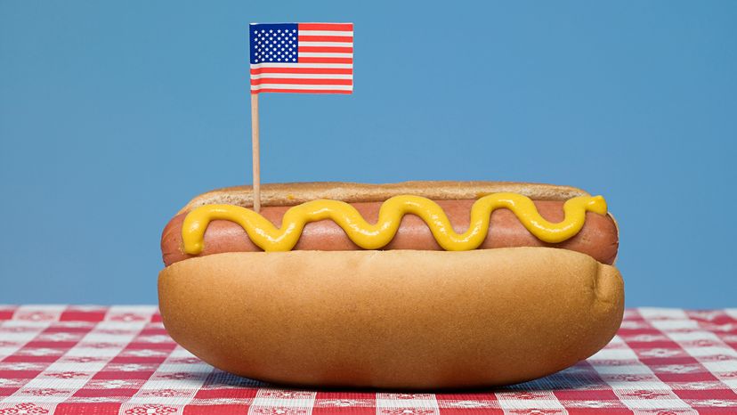A hot dog with mustard in a bun with a US flag toothpick on a red and white tablecloth