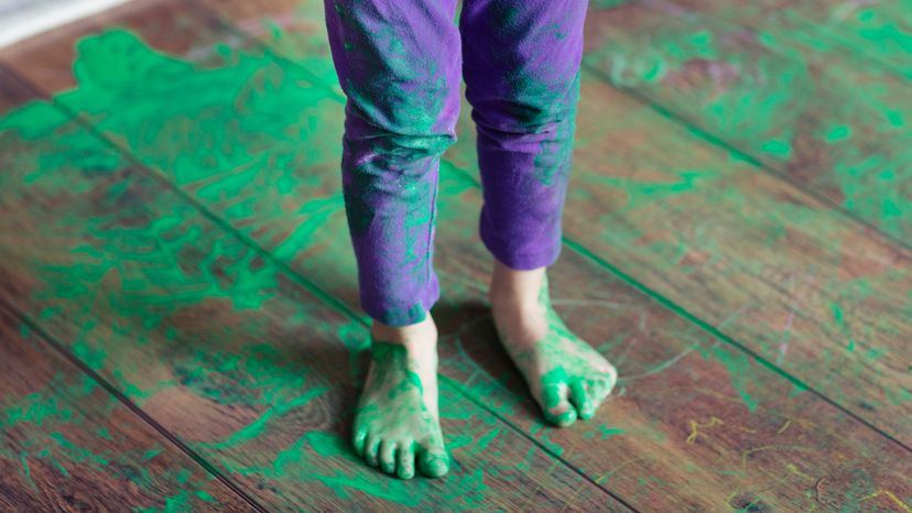 Child in purple pants standing on hardwood floor with green paint everywhere