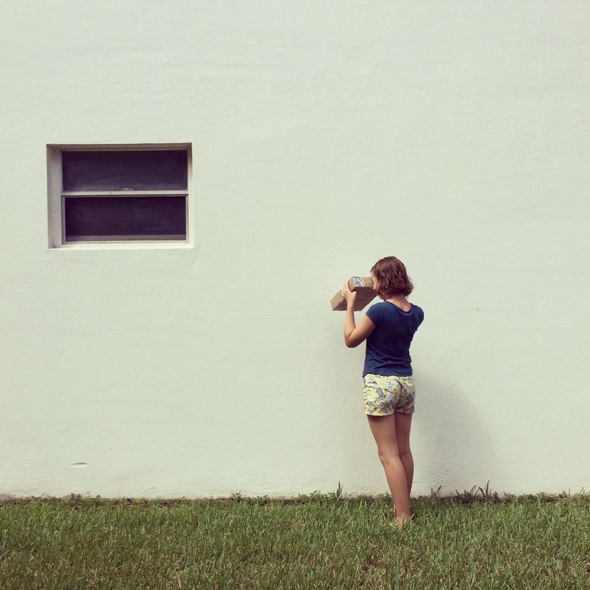 A young girl faces a white wall while looking in a homemade cardboard pinhole camera.