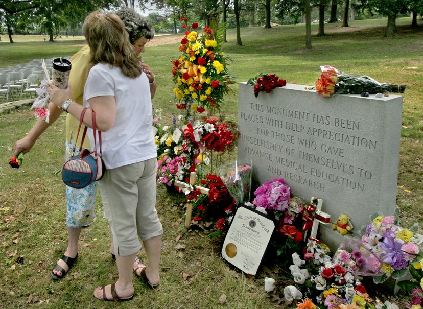 Two adults hug in front of a grave and tombstone that's covered with flowers and surrounded by grass.