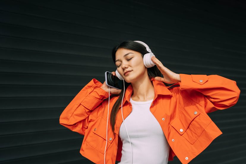 Portrait of beautiful asian dancing woman in orange jacket and white headphones on black background.