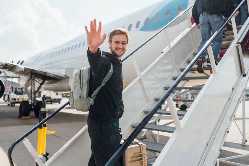 Man walking up the steps to the plane carrying his suitcase, smiling at the camera while waving.