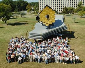 A model of the James Webb Space Telescope on the lawn at Goddard Space Center. You can gauge the size of the telescope by comparing it with the design team standing in front of it.