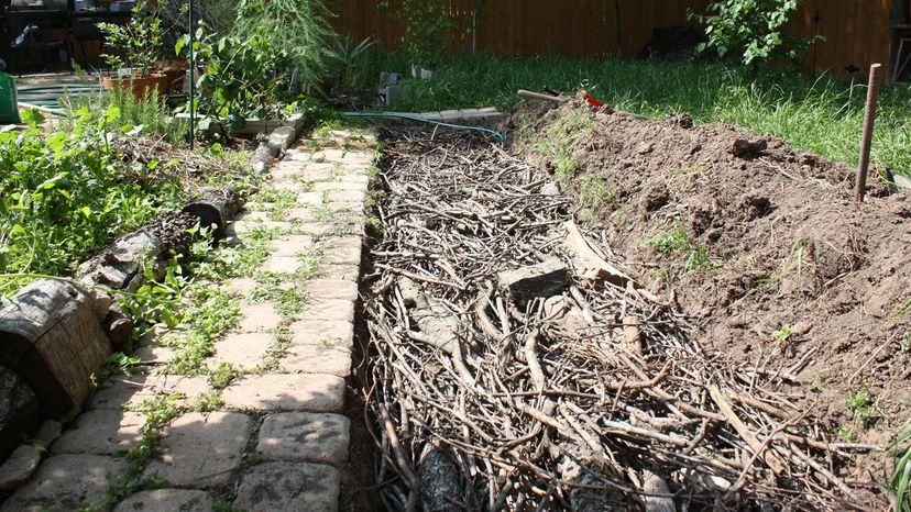 A hugelkultur bed of organic debris and branches lined up next to an old stone path.