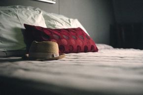 A straw bolero hat sits on a white bed with a red dotted pillow.