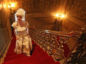 A woman in a historic dress and wig walking up a red velvet lined golden staircase.