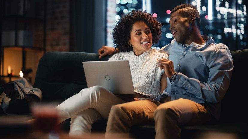 Couple in Love Use Laptop Computer, Have Fun, Laughs While Sitting on the Couch in the Cozy Loft Apartment. Couple Shopping on Internet, Using Social Media, Discussing Something.