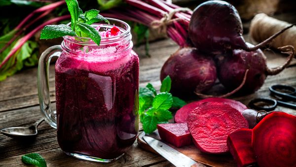 Drinking glass filled with fresh organic beet juice shot on rustic wooden kitchen table. Whole and sliced beets are all around the glass.