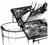 A black and white illustration of a hand using a scraper to remove paint from a table into a bucket.
