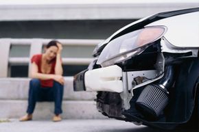 A person sits on a curb behind a wrecked car.