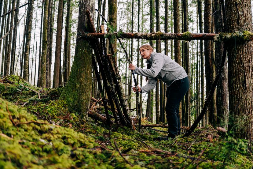 Squamish, BC, Canada - March 23, 2022: A woman places logs onto a lean-to survival shelter in a temperate rainforest on March 23, 2022.
A lean-to is a classic shelter used by survivalists in forested areas such as those in The Pacific Northwest and British ColumbiaÃ¢â‚¬â„¢s Coast Mountains.