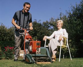 Chelsea Football Club player Ron Harris mows his lawn as his wife watches at their home in England in 1970. Mr. Harris appears to have a warm-season grass growing in his remarkable yard, possibly Bermuda.