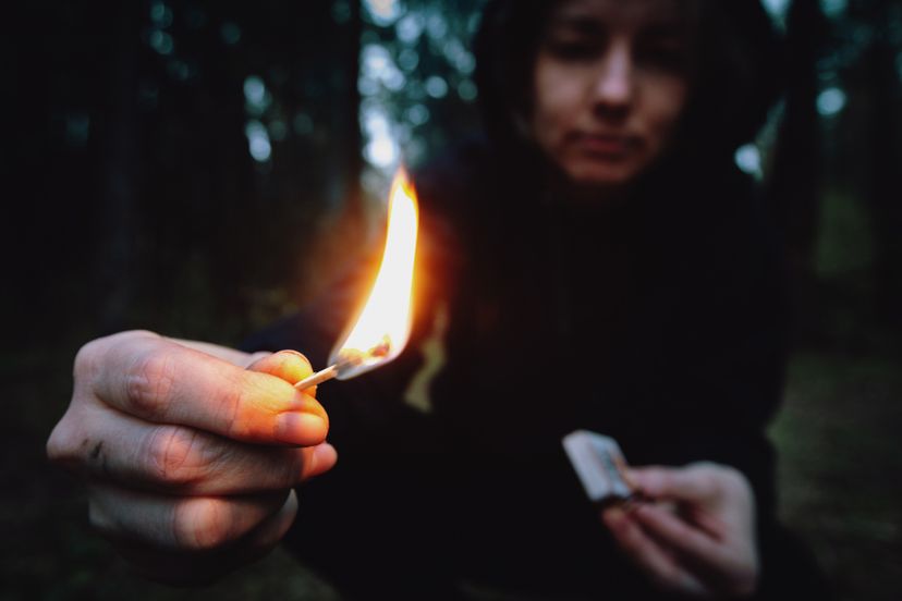 A teenager in a black hood holding a burning match in a dark forest at night. 