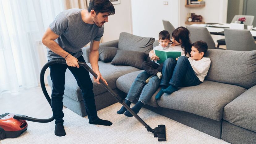 Man vacuuming while mother and boys reading book on the sofa