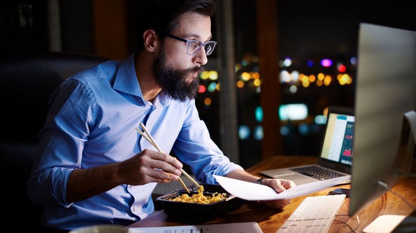 Young businessman looking at computer and eating takeaway at office desk at night