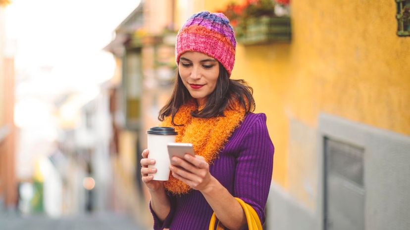 Stylish woman in city holding a cup of coffee and looking at smart phone