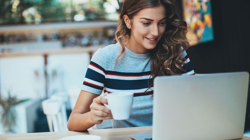 Smiling young woman sitting in cafe holding a coffee cup and looking at a laptop