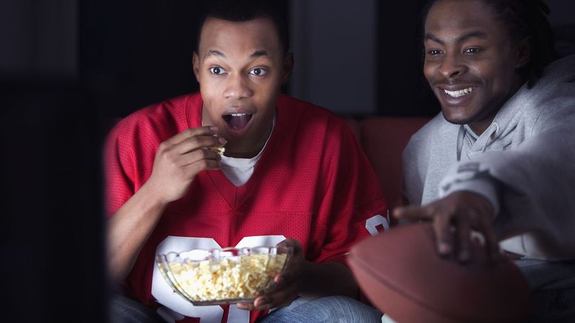 Two men holding football, watching television and eating snacks